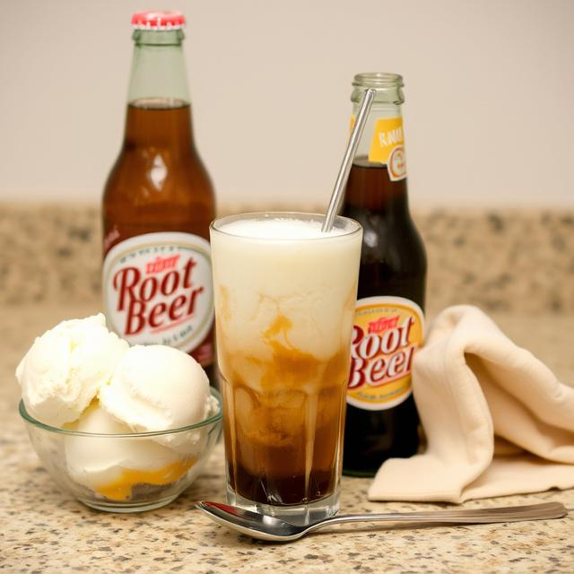 Ingredients for a root beer float — vanilla ice cream scoops, chilled root beer bottle, tall frosted glass, spoon, and straw arranged neatly on a counter.