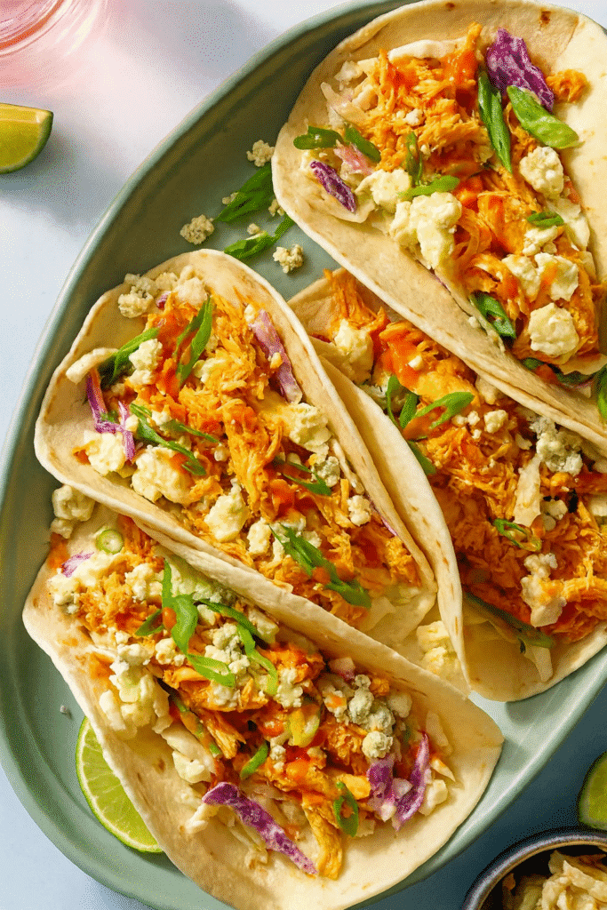 Quick Buffalo chicken tacos filled with shredded chicken, creamy coleslaw, and blue cheese crumbles arranged on a green platter over a light blue tabletop.