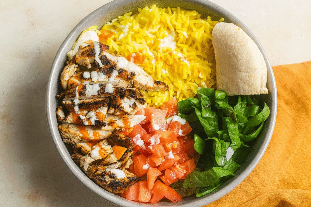 Bowl of Chicken Over Rice with White Sauce, filled with turmeric yellow rice, sliced grilled chicken drizzled with white and hot sauce, chopped tomatoes, fresh lettuce, and a wedge of pita, served on a light table with an orange napkin.