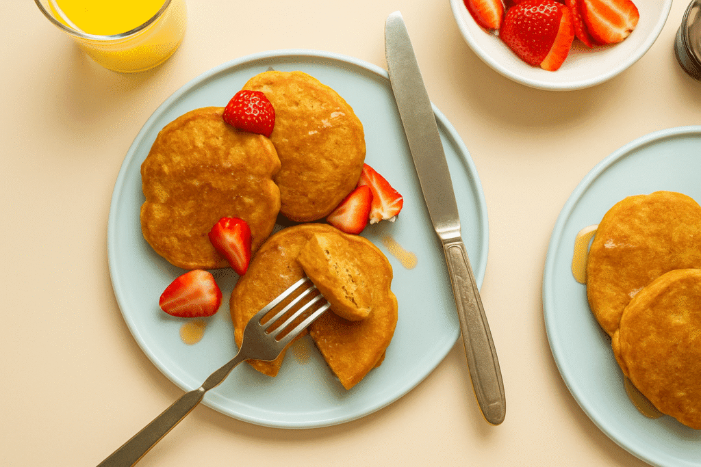Top view of 3-ingredient almond butter pancakes on a pale blue plate, with a fork cutting into one pancake, fresh strawberry pieces, syrup drizzles, a glass of orange juice, and extra strawberries on the side.