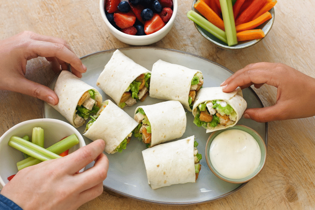 Platter of chicken Caesar wrap halves on a table with fruit and veggie sides and dipping sauce.