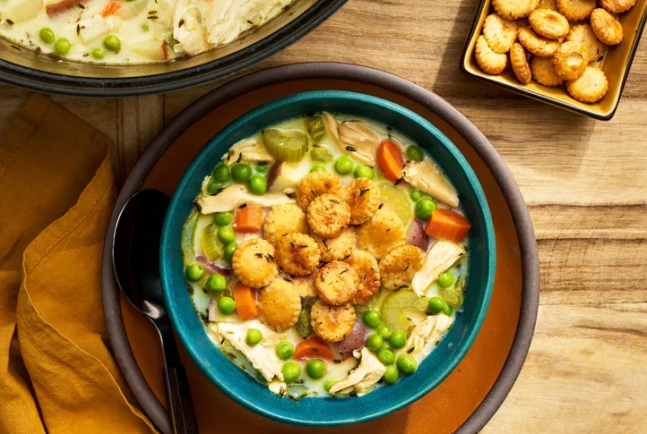 Overhead view of a teal bowl of creamy chicken pot pie soup topped with seasoned oyster crackers, peas, carrots, celery, and potatoes, set on a wooden table with a mustard napkin, spoon, and extra crackers on the side.
