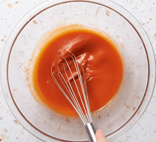 Hand whisking a bright orange Buffalo sauce in a clear glass bowl on a light countertop.