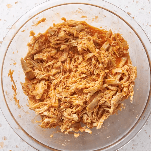 Shredded rotisserie chicken coated in Buffalo sauce in a clear glass bowl on a light countertop.