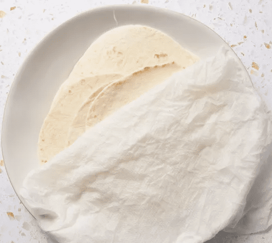 Stack of soft flour tortillas on a white plate, partially covered with a damp cloth for warming.