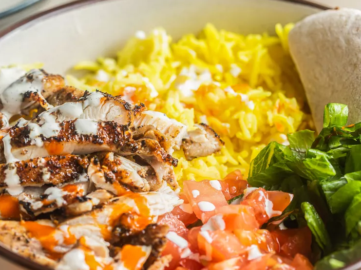 Close-up of sliced grilled chicken drizzled with white sauce and hot sauce, served over yellow turmeric rice with chopped tomatoes, lettuce, and warm pita in a bowl.