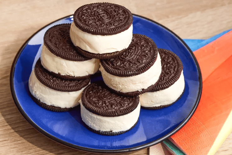 A stack of chocolate sandwich cookie ice cream sandwiches filled with vanilla ice cream arranged on a bright blue plate on a wooden table, with colorful napkins at the side.