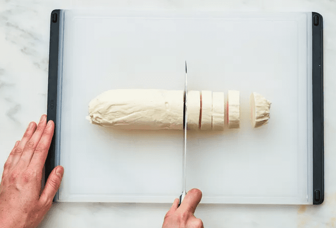 Hands slicing a chilled log of vanilla ice cream into even rounds on a white cutting board, preparing pieces for easy two ingredient ice cream sandwiches.