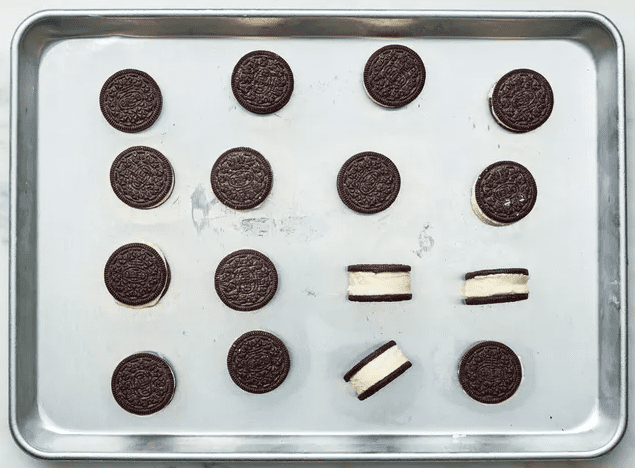 Chocolate sandwich cookies arranged in rows on a metal baking sheet, with a few cookies filled with vanilla ice cream to form mini ice cream sandwiches.