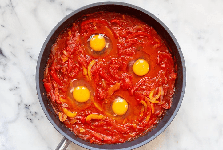 Thickened tomato and pepper sauce with a spoon making small wells for eggs in a skillet.
