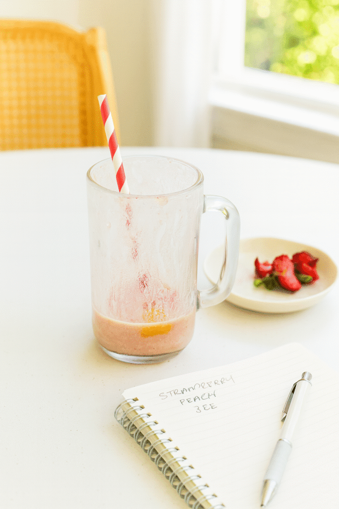 Nearly empty Strawberry Peach Smoothie glass beside a recipe notebook and pen on a small table