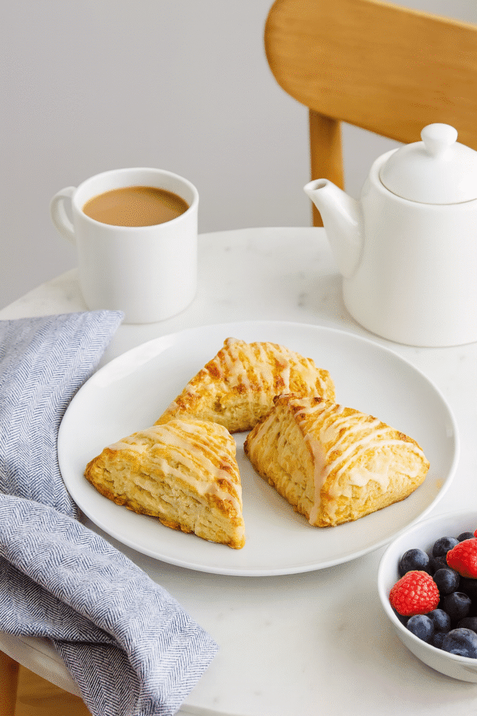 Cozy brunch table with a white plate of three glazed lemon scones, a cup of coffee, a white teapot, a bowl of fresh berries, and a folded cloth napkin.