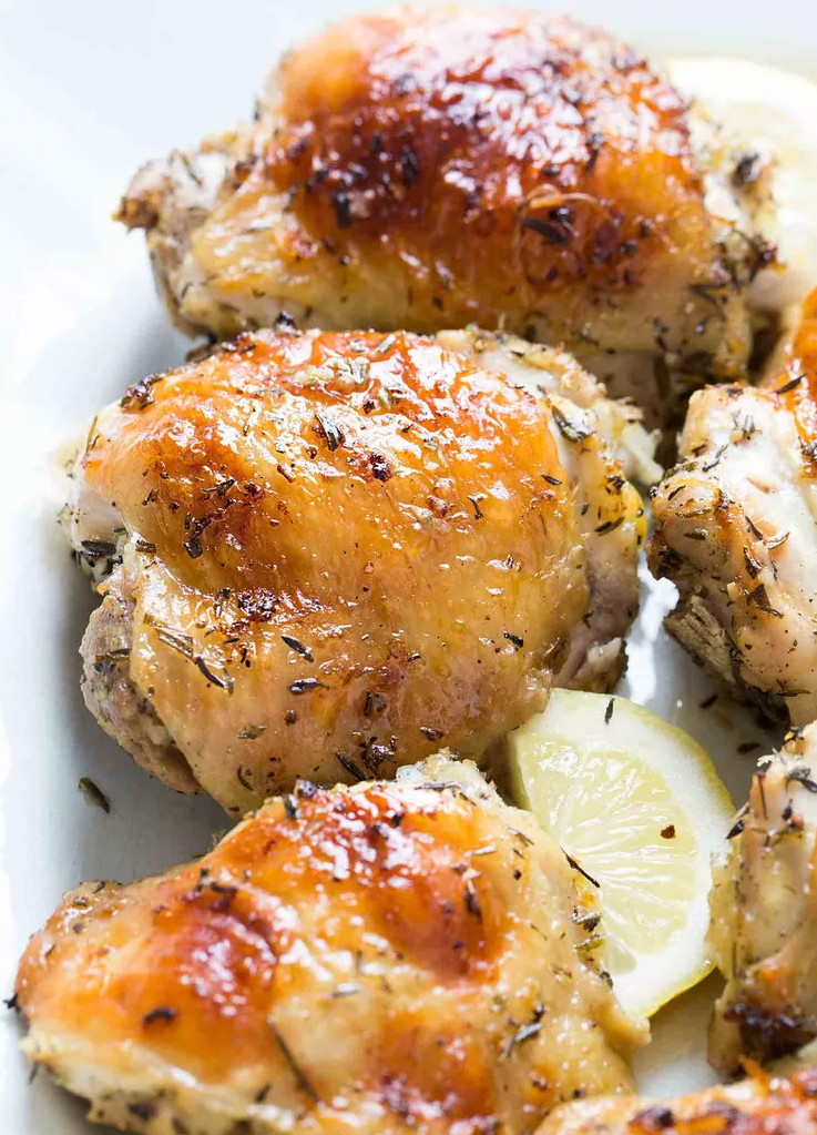 Close up of oven baked lemon chicken thighs with crispy golden skin speckled with herbs and a lemon wedge tucked between the pieces