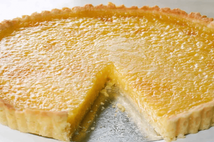 Bright hero shot of a whole lemon tart on a light surface, one slice removed, lemon zest curls and a knife nearby