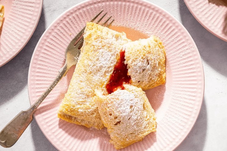 Close up of two powdered sugar coated toast pies on a pink plate, one broken open to show bright red jam filling beside a fork.