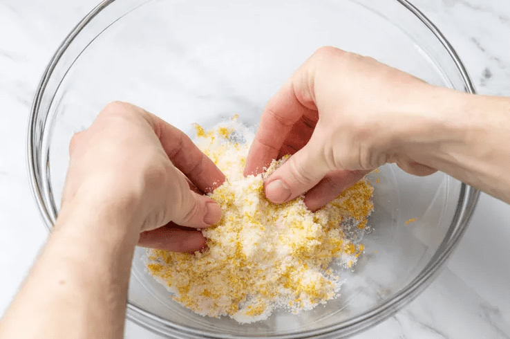 Hands rubbing fresh lemon zest into granulated sugar in a clear glass bowl to combine the dry ingredients for bright lemon scones.