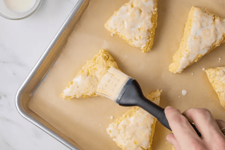Unbaked lemon scone wedges on a parchment-lined baking sheet, with a hand using a pastry brush to coat the tops with cream.