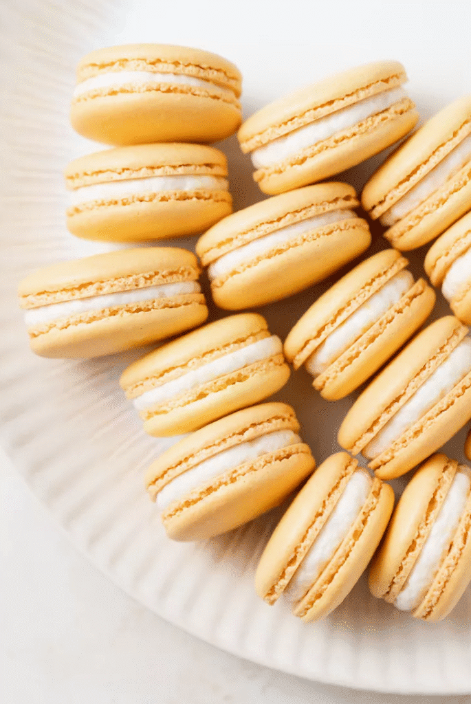 Overhead view of bright yellow lemon macarons arranged in a loose circle on a white plate, each filled with a creamy lemon buttercream.