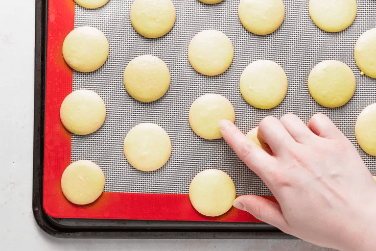 Rested lemon macaron shells on a tray with a finger lightly testing the dry surface.