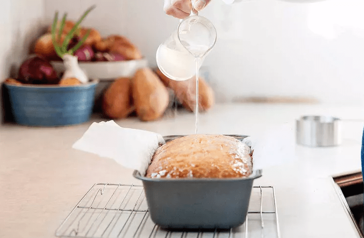 Hand pouring clear lemon syrup over warm lemon pound cake with small holes poked across the surface